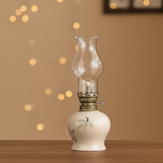 A clear glass oil lamp with a white ceramic base and brass collar sits beside a small clear plastic bottle with a white cap and printed label, on a wooden surface against a tan background. A black text bar at the bottom reads: Diameter ≈ 3.