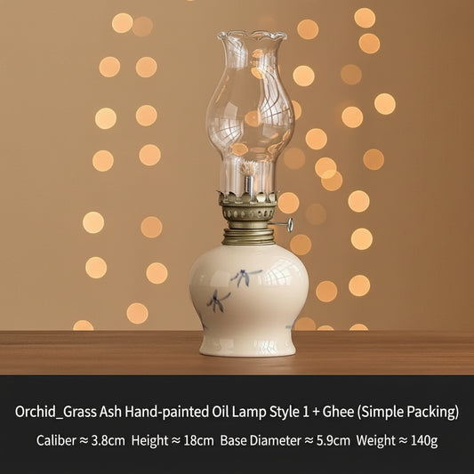 A hand-painted oil lamp in a cream-colored ceramic base with a clear glass chimney and brass burner, shown next to a small bottle of ghee on a wooden surface against a tan background. A black text banner at the bottom displays product details.